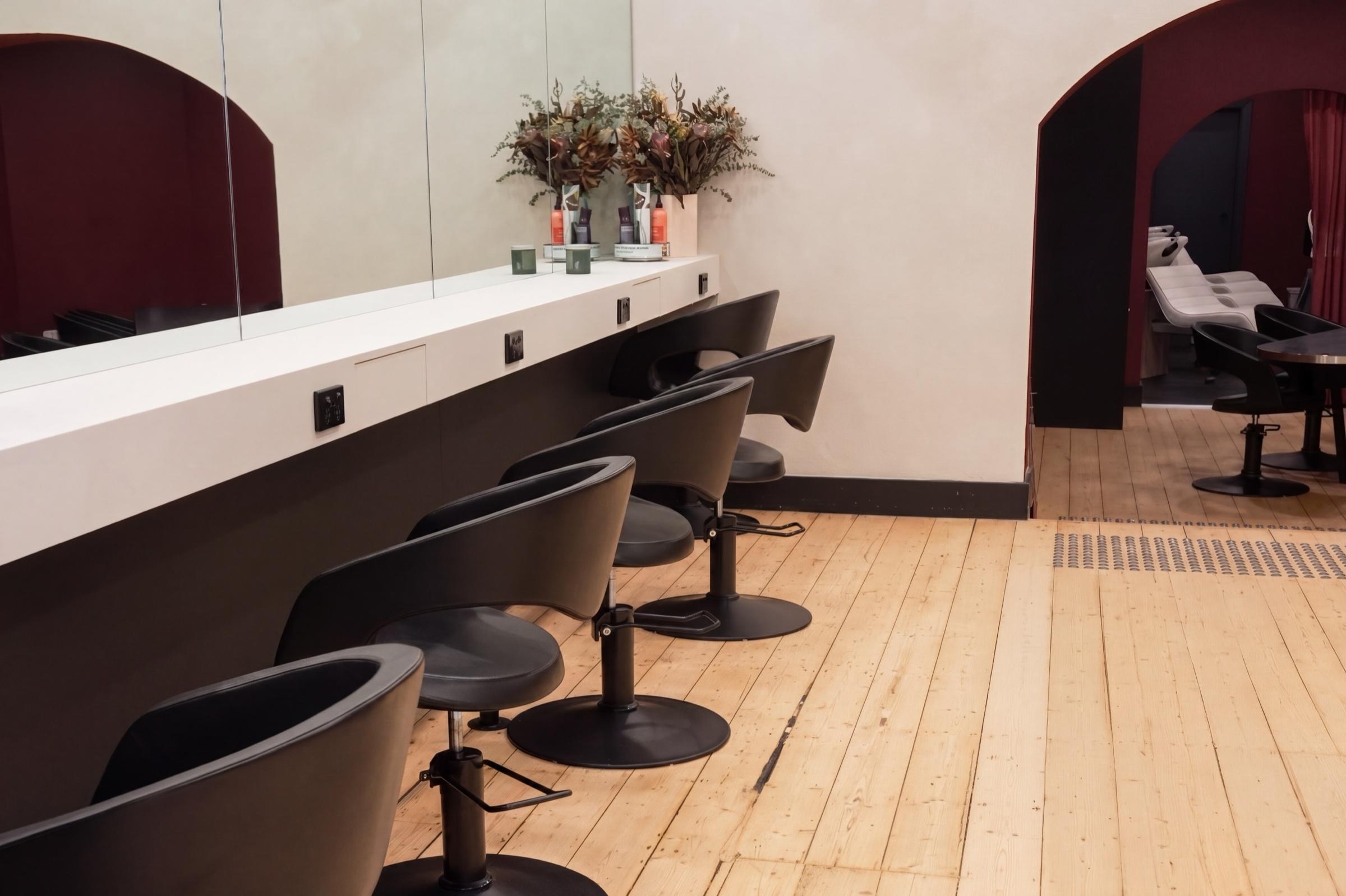 Black styling chairs receding along the mirrored commune floor, blond timber underfoot, the deep red arch of the wash sanctuary open at the right